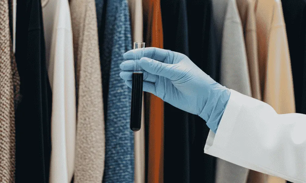 Gloved hand holding test tube with dark liquid in front of racks of clothing, indicating textile chemical analysis in progress. - chemical testing of textiles 2

