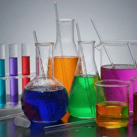 Laboratory glassware with colorful chemical solutions in flasks and beakers arranged against a neutral background. -Textile Pre-Treatment Chemical Suppliers. -1