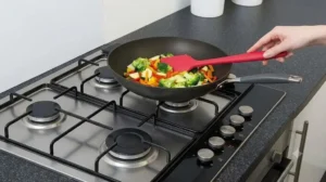 A person uses a red silicone spatula to stir-fry vegetables in a pan on a gas stovetop.