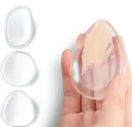 Silicone makeup blender held in hand with foundation applied to its surface, alongside three clear silicone makeup blenders on a off white background. - silicone makeup blender 2