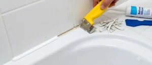 A person using a yellow sealant scraper tool to remove old white silicone caulking from a clean, white-tiled bathroom bathtub joint.